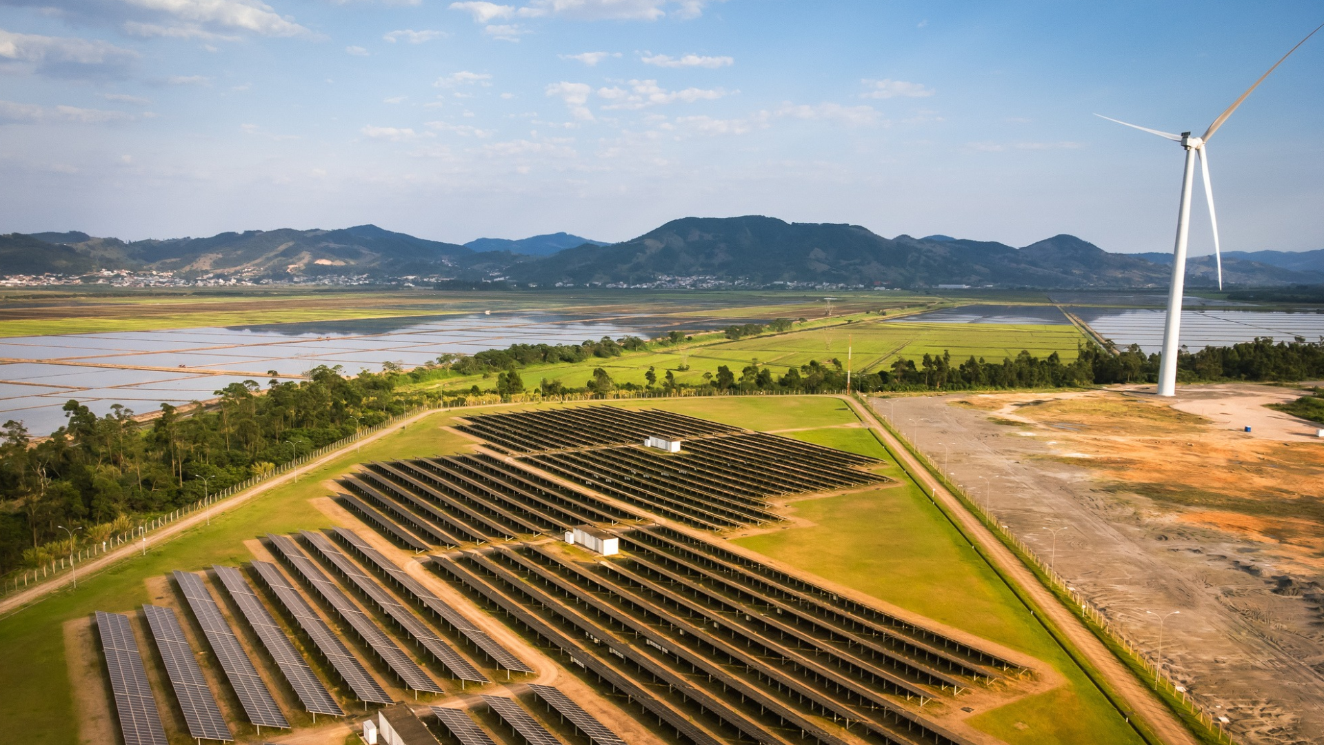 Image of wind turbine and solar panels in a lush green field with mountains in the background. 