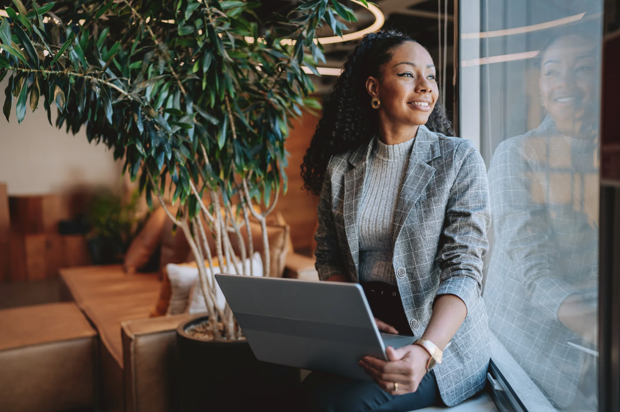 Lendable MSME Fintech Credit Fund II géré par Lendable Inc : Photo d’une femme d’affaires tenant un ordinateur portable et regardant par la fenêtre. Une plante et un sofa sont présents en arrière-plan.
