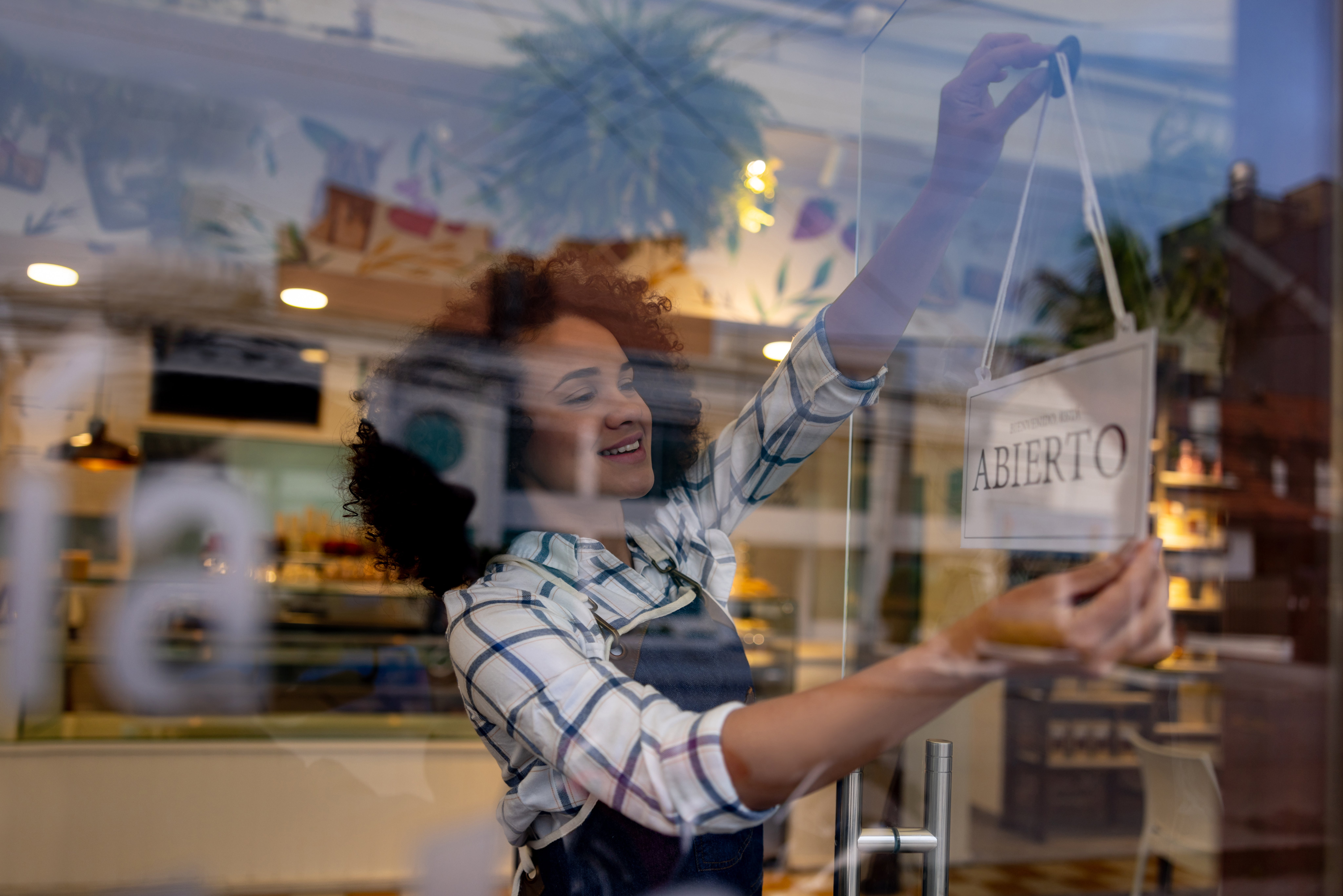 A smiling waitress hanging an “OPEN” sign in Spanish at a café.